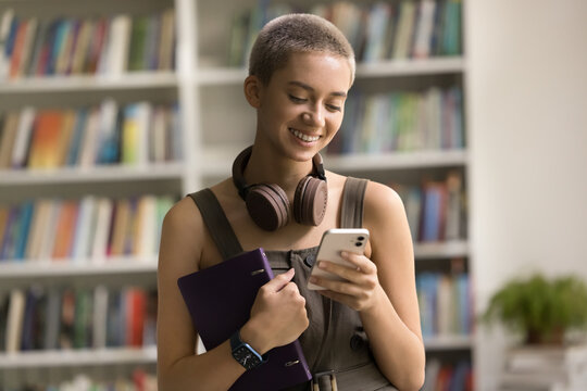 Pretty student girl using modern smartphone in library, chatting with schoolmate, check online schedule using comfortable mobile application, read messages, share sms with friends via social media app