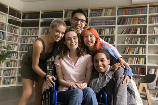 Portrait of five diverse schoolmates in library. Girl with physical disability sit in wheelchair posing for cam surrounded by friendly schoolmates, smile, express warm relation. Equality, friendship