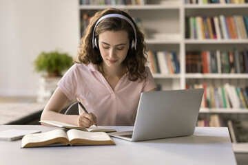 Attractive teenage student girl wear headphones studying in library, sit at desk with laptop, make...