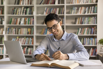 Smiling intelligent Indian student guy in glasses sit at desk, make exercise, writes in copybook,...