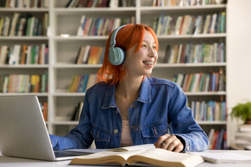 Red-head student girl listen audio course studying on-line sit at desk with laptop and textbooks in...