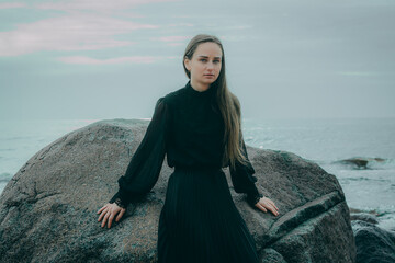 A woman in dark clothes is sitting on a rock on the shore