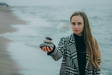 woman is holding a pyramid of stones in her hand, waves are crashing behind her. Black dress plaid coat. Cloudy sky.