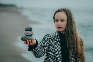 woman is holding a pyramid of stones in her hand, waves are crashing behind her. Black dress plaid...