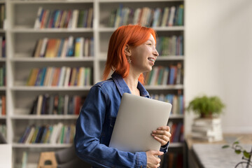Happy red-headed girl student holding laptop standing in college library, smiling, looks away,...