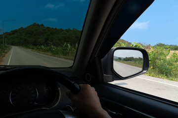 View from the inside of a car. View from the driver's seat. Driving on the asphalt road with nature of tree and mountain beside road. Under blue sky in the day time.