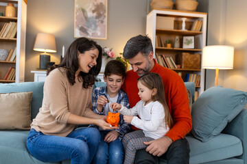 Happy family cheerful mother and father with kids smiling and putting coins into piggy bank while sitting on sofa at home