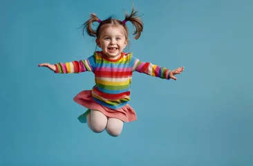 Fotobehang A little girl in striped colorful dress with pigtails hair jumping up and smiling on blue background © Kien