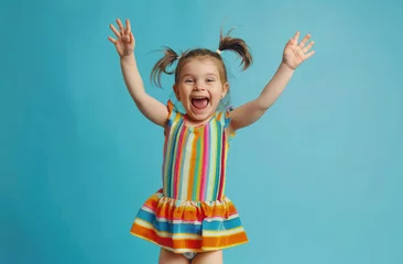 Fotobehang A little girl in striped colorful dress with pigtails hair jumping up and smiling on blue background © Kien