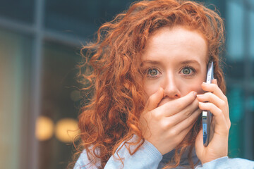 Portrait of beautiful shocked young woman with curly red hair talking on the mobile as she getting...
