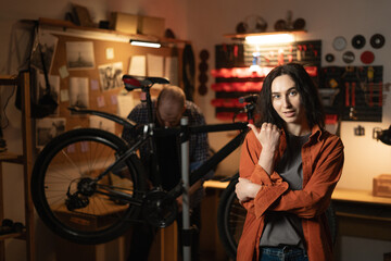 Male mechanic working in garage repairing bicycle, woman standing in foreground pointing with finger at space for text