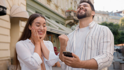 Caucasian happy couple enjoy excited man woman shocked lucky success victory mobile phone...