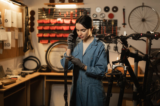 Female mechanic takes order by mobile phone standing in bicycle workshop near bike. Mechanic talking on mobile phone after fixing bike.