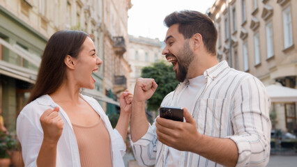 Caucasian couple happy woman man rejoices winning victory lucky expression together joy using mobile phone cellphone high-five exclamation success achievement emotions delighted triumph city outside