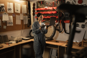 Young female mechanic in bike workshop working and researches information on his laptop.