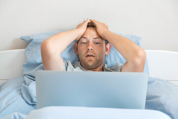 Shocked man lying in the bed and working on a laptop. Amazed Caucasian male freelancer surfing and browsing internet, working online from home and stunned with bad news. Distance job