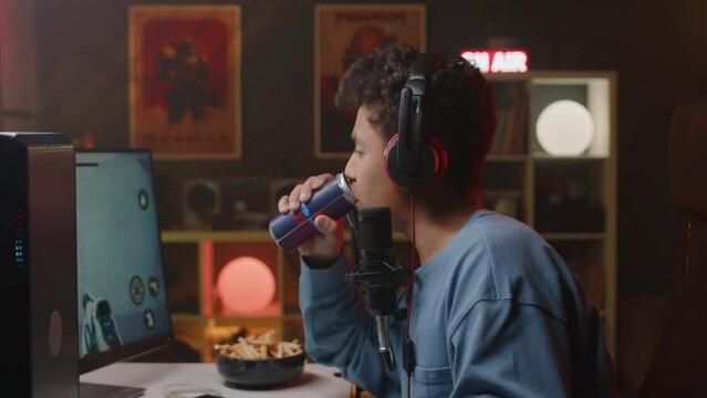 Side view waist up shot of Biracial teenage boy in headset playing online first person shooter game on personal computer in neon light room in apartment at nighttime