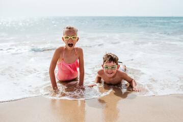 Siblings playing on beach, lying in water, having fun. Smilling girl and boy in swimsuits, swimming googles on sandy beach of Canary islands. Concept of family beach summer vacation with kids.