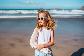 Girl with broken arm on beach. Young blonde girl has arm cast, injured during family vacation in holiday resort. Concept of beach summer vacation with kids.