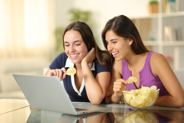 Two happy friends watching movie on laptop eating snack