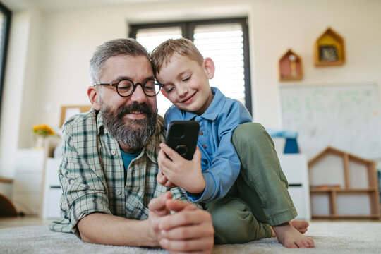Little boy watching cartoon movie on smartphone with father, lying on floor in kids room. Dad explaining technology to son, digital literacy for kids.