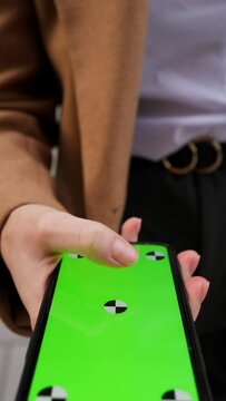 Vertical view of closeup hands of young woman holding and touching phone with green screen vertically in the street on go 