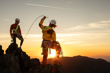 Two people are standing on a mountain, one of them holding a rope. The sun is setting in the...