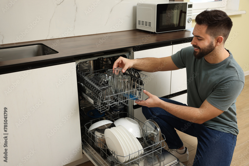 Sticker smiling man loading dishwasher with glasses in kitchen