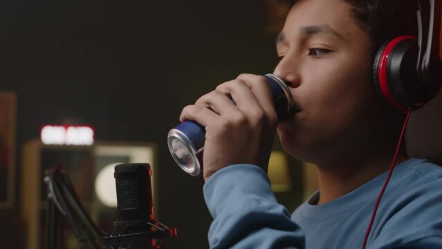 Tilting up shot of young Biracial male gamer opening can with energy drink and taking sip while doing live game streaming and playing at neon light room at night