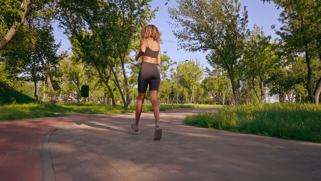 A woman in athletic attire is jogging at a steady pace down a concrete sidewalk in a park, surrounded by trees and greenery. She is focused and determined in her movements, with her arms swinging in