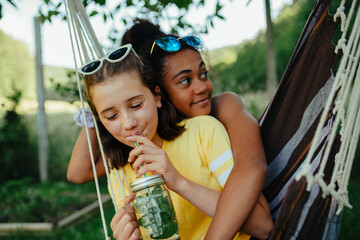 Teenage girls friends outdoors in garden, swinging on swing, drinking lemonade from jar glass, during warm summer evening.