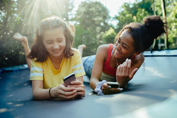 Teenager girls friends spending time outdoors in garden, laughing. Lying on trampoline, scrolling on smartphone, social media.