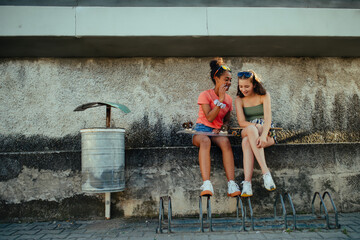 Teenager girl best friends with skateboards spending time outdoors in city during warm summer holiday day. Sitting on concrete wall, talking, sharing secret.