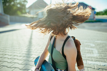 Teenager girl with skateboard spending time outdoors in city during warm summer holiday day. Flying hair, spinning head.