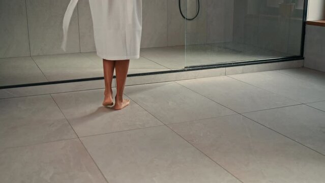 A person wearing a bathrobe walks towards a shower, preparing to bathe. The individual approaches the shower area with intent and purpose, ready to begin their washing routine. The tiled bathroom