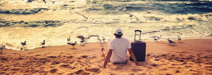 A human sits on the beach in summer with a travel suitcase. The man looks at the sea. Seagulls fly over the beach