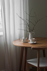 A cup of tea, a teapot, branches in a jug on a round wooden table in the living room - 785060371