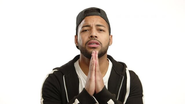 A young man with clasped hands appeals earnestly, his facial expression showing genuine hope and a request for help or understanding, against a blue background. Camera 8K RAW. 