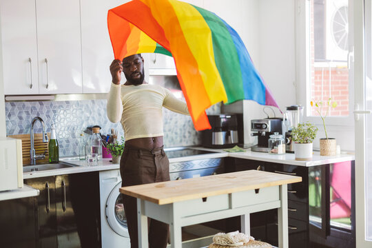 Young non-binary person holding rainbow flag near table at home