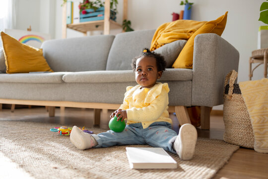 Cute girl sitting with toy and book on carpet in living room at home