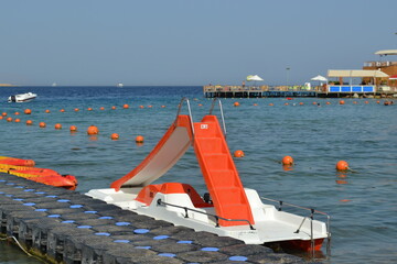 A white catamaran with an orange slide and seats is parked near a plastic pier in Egypt. In the sea...