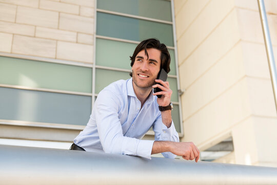 Smiling young businessman talking on smart phone leaning at railing