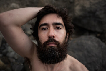 A scruffy, bearded man with messy hair sits on a rocky beach and gazes at the camera. He lifts up...