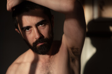 Obraz premium Shirtless bearded man with arm raised, looking directly at the camera. Lighting from the window sets a dramatic tone, with a heavy shadow across his face.