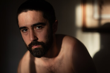 Shirtless man with beard in a dimly lit room. The light leaves dramatic shadows on his body and the background. He looks directly at the camera.