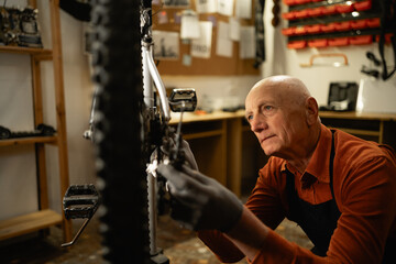 Old male bike shop mechanic repairing bicycle wheel in workshop or garage