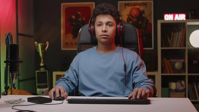 Medium shot of Biracial gen z pro gamer in professional headset with mic operating gaming mouse and keyboard while playing online video game on computer and live streaming
