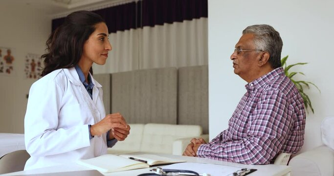 Happy young Latin practitioner woman meeting with elderly Indian patient, listening to health complaints, nodding, talking to mature man, smiling, giving consultation for checkup
