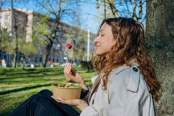 A young woman enjoys a fresh salad and raspberry outdoors, relaxing against a tree in a sunny urban park. 