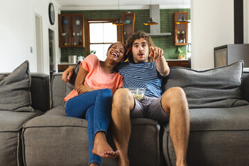 Diverse couple enjoying time on sofa, pointing at something, watching television or movie at home